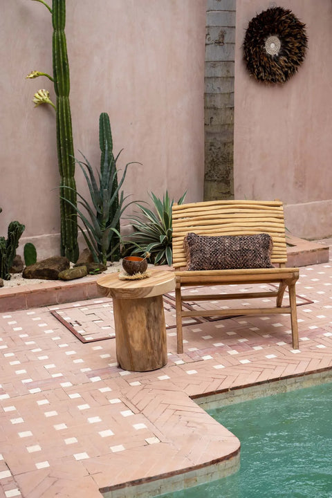 A serene outdoor patio showcases a teakwood chair beside a small round table. The area features unique design elements like potted cacti against a stucco wall and a decorative sunburst wall hanging, all overlooking a turquoise pool to create a tranquil ambiance.