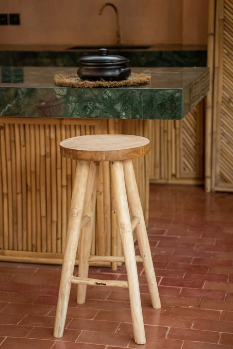 A teak wood stool stands on a red-tiled floor in front of a bamboo-paneled kitchen counter with a green marble top. A black pot rests on the counter. This setting, reminiscent of an outdoor bar, exudes a natural, rustic aesthetic.