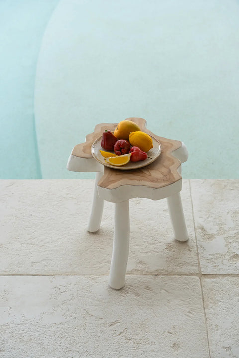 A small teak wood side table by the poolside holds a plate with lemons, strawberries, and slices of lemon. The background showcases the edge of a serene, light blue swimming pool and textured stone tiles.