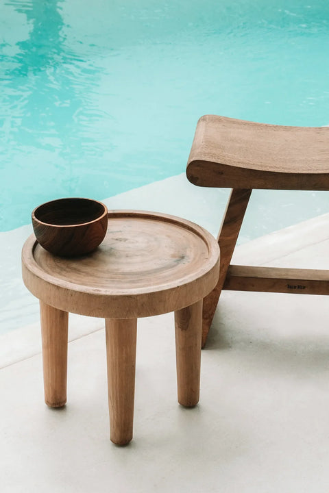 A chic wooden stool and a suar wood side table with a bowl on top are placed near a turquoise swimming pool. The minimalist design exudes a natural style, creating a calm and serene setting that evokes relaxation by the poolside.