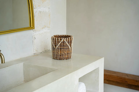 A rattan basket exudes natural elegance on a stone countertop in a minimalist bathroom. The countertop hosts a rectangular sink, complemented by an off-white wall and wooden shelf. A brass-colored mirror frame peeks through, adding to the serene ambiance.