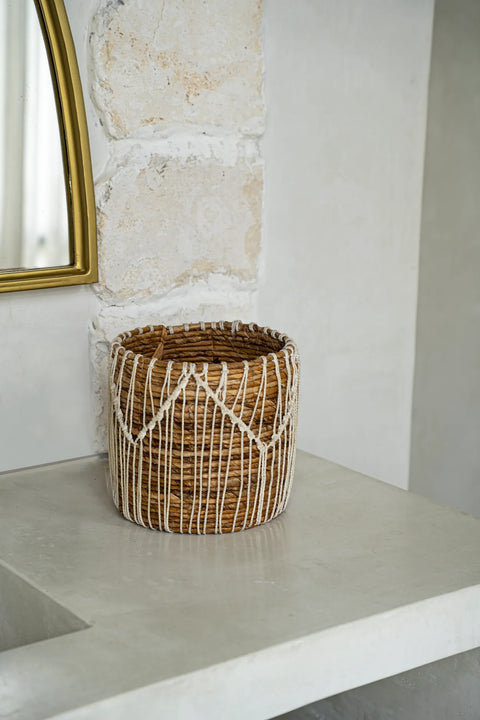 A Macra-Mazing Basket, featuring intricate white rope patterns in a macramé design, sits on a light-colored stone shelf. A portion of a mirror with a gold frame is visible on the left side, set against a textured wall.