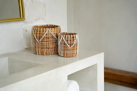 Two woven Macra-Mazing Baskets, one taller than the other, sit on a smooth, light-colored surface. They boast a natural, rustic appearance with shades of brown and subtle banana leaf accents. A golden-framed mirror peeks from the left, while a wooden shelf stands to the right.