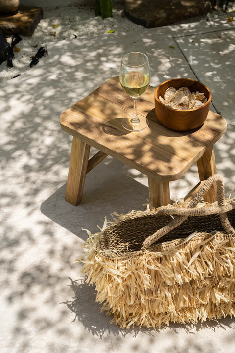 A rustic stool crafted from reclaimed teak wood holds a glass of white wine and a small bowl of chips. A straw bag rests nearby, embodying eco-conscious charm. Dappled sunlight creates leaf patterns on the ground, enhancing the serene ambiance.