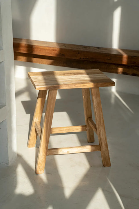 Sunlight streams through a window, illuminating the Oriki Stool crafted from reclaimed teak wood. The wooden stool, with its rectangular seat, stands gracefully on a light-colored floor, casting intricate shadows that dance around it.