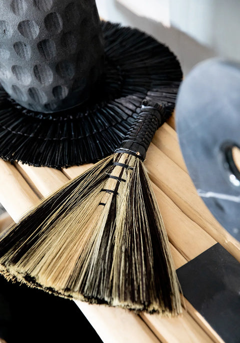 A close-up of a decorative straw broom, reminiscent of boho style, with a black wrapped handle, resting on a light wooden surface. In the background, a textured wide-brimmed black hat adds to the aesthetic charm.