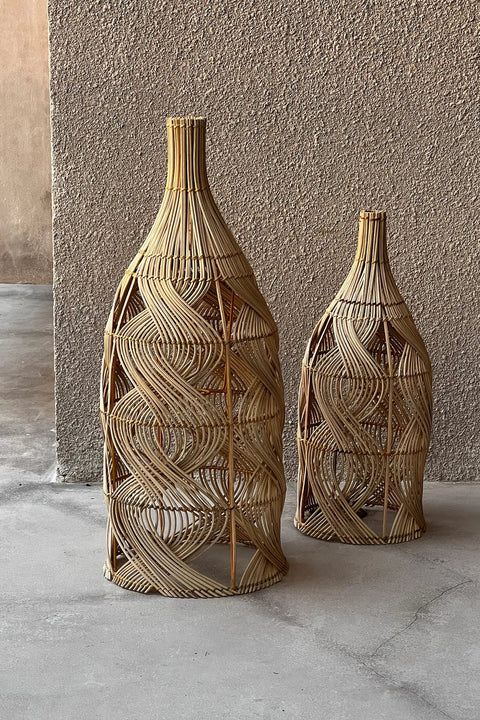 Two decorative wicker bottle covers of different sizes stand on a concrete floor against a textured wall. The woven patterns create an intricate, swirling design, reminiscent of an artisanal rattan lamp that adds a touch of elegance to any space.