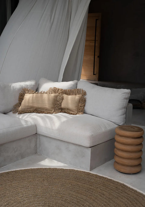 A cozy living space featuring a white sectional sofa with beige fringed pillows. A textured round rug, woven with jute, is in front, and a unique cylindrical side table is beside the sofa. Soft, airy curtains in the background add a serene touch to the soft furnishings.