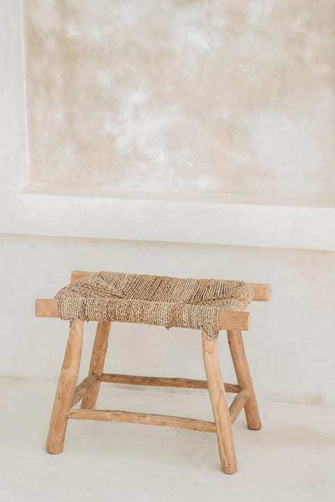 A rustic teak wood stool with a seagrass woven seat is placed against a neutral-toned, textured wall. The minimalist setting highlights the stool's natural wood and woven design, creating an earthy and serene boho style atmosphere.