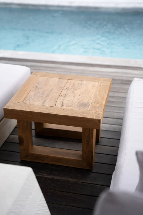 A wooden square coffee table made from reclaimed teak wood is placed on a deck beside a pool. The table boasts a simple, rustic design and is surrounded by white cushioned seating, creating a serene and sustainable minimalist outdoor setting.