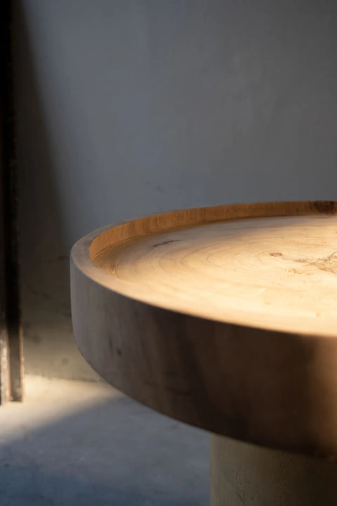 A close-up of a round suar wood side table with a smooth, natural finish. Soft, warm light highlights the timeless piece's grain and texture, casting shadows on a gray background. The scene conveys a calm and minimalist aesthetic.