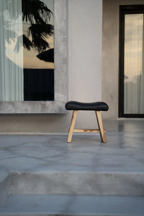 A minimalist suar wood stool with a matte leather cushion sits on a smooth concrete patio in front of a modern building. A large window reflects a silhouetted tree, and a door is partially visible on the right, embodying natural classic style.