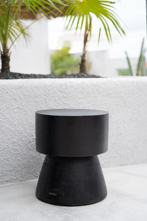 A sleek, modern Warmi stool with a cylindrical seat and conical base is crafted from black-coated suar wood. It sits elegantly on a light-colored patio, while palm plants add greenery against a white textured wall. The setting exudes futuristic design with its minimalistic and stylish appeal.