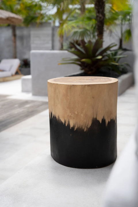 A wooden stool with a dip-dyed natural wood top and black painted base stands outdoors on a light concrete patio. In the background, there are tropical plants and a lounge chair with an umbrella, adding to the charm of chunky furniture in the serene setting.