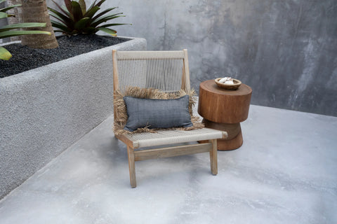 A wooden chair with a gray cushion and woven fringe detail is placed next to a rounded wooden side table. The setting is an outdoor patio with concrete flooring and a planter with succulents in the background.