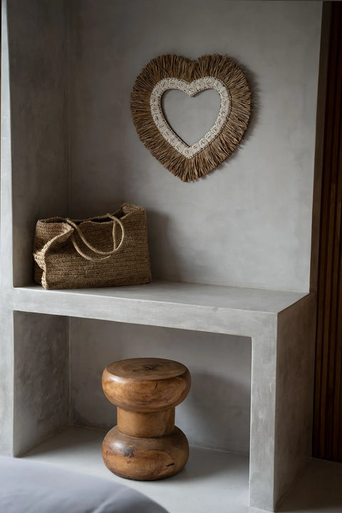 A Wayra stool crafted from suar wood sits under a minimalist concrete shelf, which holds a woven straw bag. Above, a heart-shaped decorative mirror with an organic shape and textured straw frame hangs on the wall.