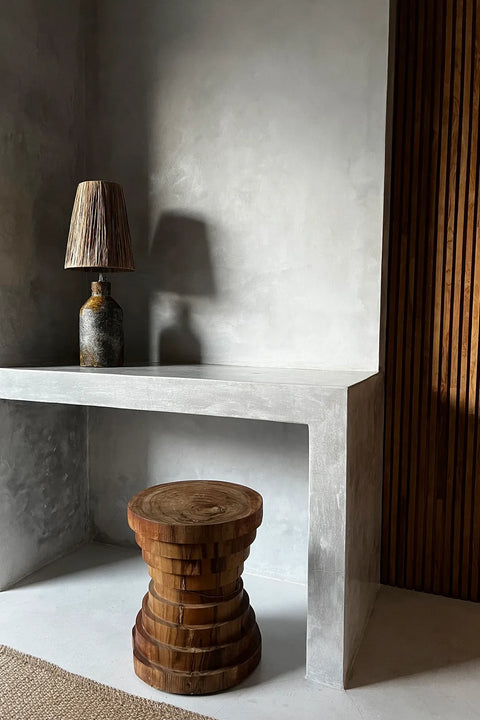 Minimalist interior featuring a concrete desk with a rustic lamp and an Indonesian suar wood stool. The room has textured gray walls and a section of vertical wooden slats, embodying contemporary design and creating a warm, inviting atmosphere.