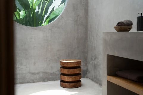 The minimalist bathroom features gray concrete walls and a round window framing vibrant green leaves. An elegant Massaka stool crafted from Suar wood with a unique layered design complements the space. On the countertop rest rolled towels and a sleek soap dispenser.