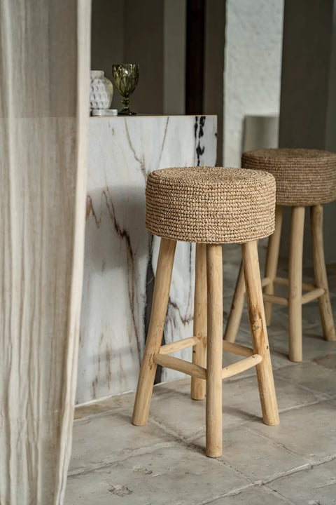 A cozy interior features two raffia bar stools with woven seats in front of a marble counter. A sheer curtain drapes partially across the scene, and a glass with decorative texture is placed on the counter, highlighting the charm of sustainable teak wood craftsmanship.