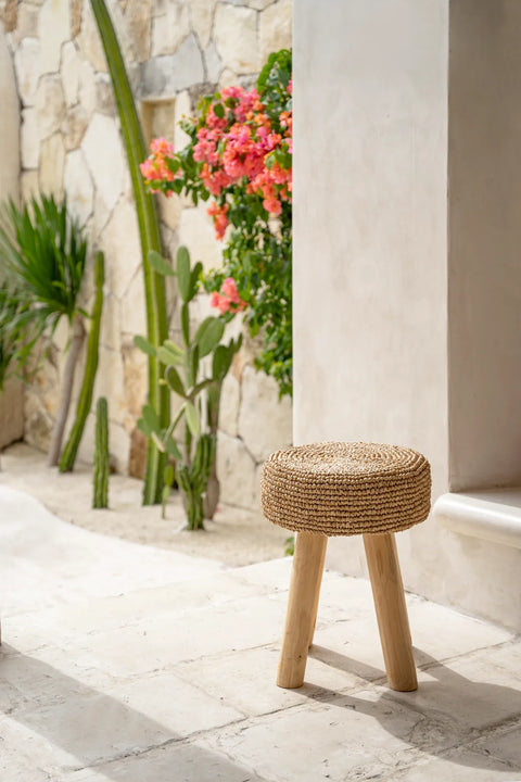 A small, woven raffia stool with three wooden legs is placed on a tiled patio. In the background, a stone wall with tall cacti and lush pink and orange flowers adds a vibrant touch to the serene outdoor setting.