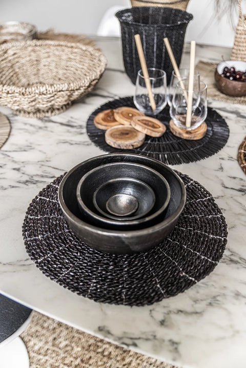 A neatly arranged table setting features unique tableware, including nested black bowls on a woven placemat, wooden coasters, two glasses with wooden straws, and a basket in the background. The light marble-patterned table surface adds an elegant touch.