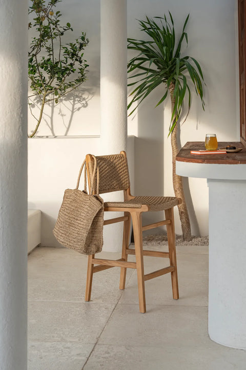 A bar stool crafted from ecological teak wood, with a woven seat and backrest, stands by the counter holding a woven bag. A glass of juice rests nearby. In the background, a tall plant and small tree cast their shadows gracefully.