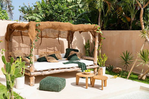 A cozy outdoor seating area features a wooden daybed with green and brown cushions under a woven canopy made of coconut fiber. Two small wooden tables with a bowl of apples sit nearby, surrounded by lush tropical plants and a white stucco wall, adding an organic touch.