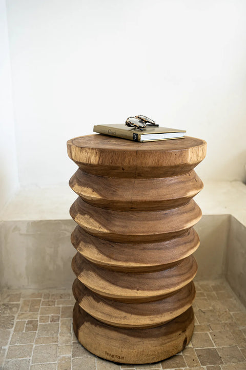 A round wooden side table, expertly handmade from suar wood, features a stacked, ridged design and rests on a tiled floor. On top, two books and a pair of glasses await. In the background, a white wall with a built-in bench completes the scene.