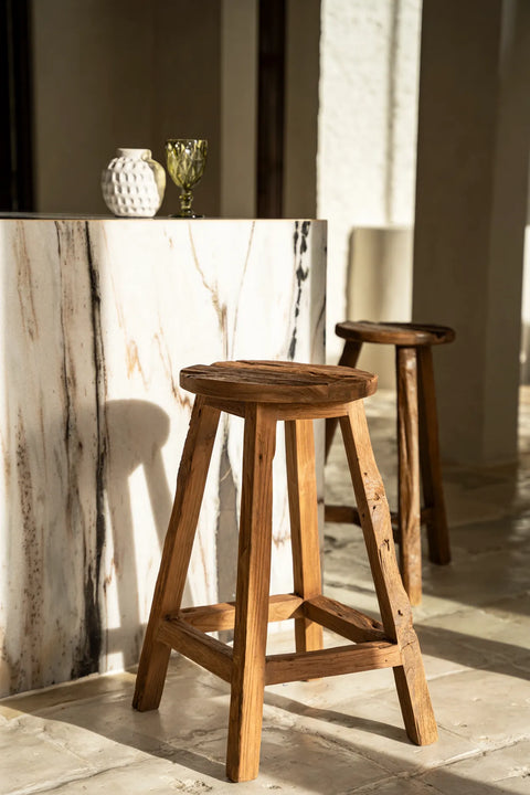 Rustic bar stools crafted from reclaimed teak wood stand by a marble bar with a vase and a green goblet on top, bathed in soft sunlight. The setting suggests a minimalist and elegant interior space, offering both style and comfortable seating.