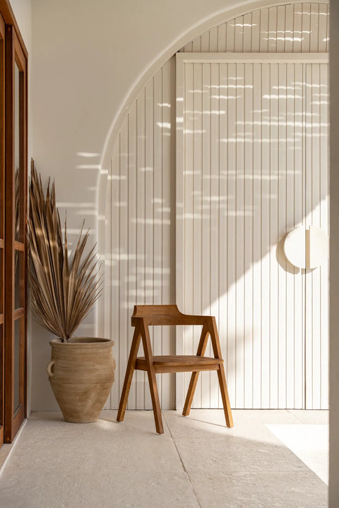 Minimalist interior with a Nihi Sumba chair, a large clay pot holding dried palm leaves, and a white paneled wall featuring an arched doorway. Soft light creates patterns on the wall, adding warmth to the serene setting, enhanced by the charm of teak wood accents.