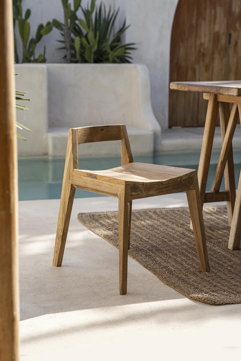 A reclaimed teak dining chair rests on a textured rug beside a matching table. Set on a patio with a smooth floor, the scene is framed by desert plants and a curved wall, with a small pool glistening in natural light. This eco-friendly setting exudes tranquility and style.