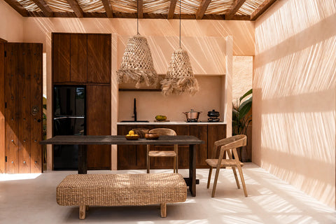 A rustic kitchen features wooden cabinets and a dark dining table with chairs. Two Abaca Jelly Fish Pendant lights hang from a wooden ceiling, casting coastal charm throughout. Sunlight filters in, creating shadows on peach walls. A bench and fruit bowl add to the natural decor.