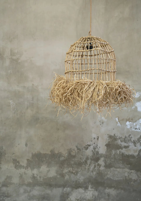 A rustic Casita pendant made of woven natural fibers and adorned with straw-like fringe evokes the essence of a tropical paradise, set against a textured, weathered concrete wall.