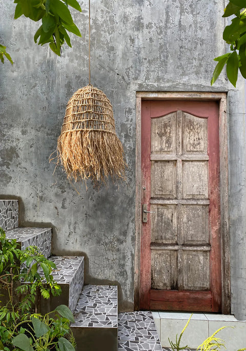 A rustic wooden door nestles in a weathered stone wall, with a Casita pendant made of woven straw casting a warm glow. Mosaic-tiled steps lead up to this tropical paradise, surrounded by lush green plants and exotic leaves.