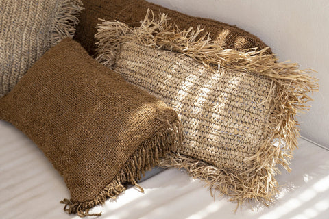 A cozy arrangement of handmade textile pillows in earthy tones with fringe details, resting against a white wall. Soft sunlight creates gentle patterns on the woven surfaces, enhancing the boho vibe.