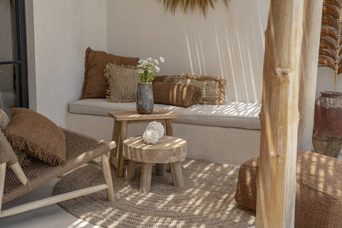 A cozy, rustic patio featuring a cushioned bench adorned with hand-dyed, brown pillows exudes a subtle boho vibe. A small wooden table and wicker chair complete the scene, with a vase of flowers and decorative dried plants enhancing the warm, natural atmosphere.