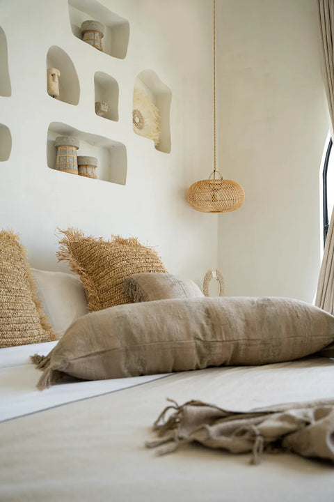 A cozy bedroom with a neutral color palette features a bed adorned with beige pillows and a velvet cushion for added luxury. Built-in wall shelves hold decorative items, and a woven pendant light hangs above, introducing a boho vibe to the serene space.