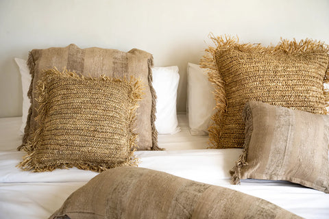 A bed with white sheets is adorned with decorative cushions, including two rectangular beige pillows with striped patterns and two square fringed pillows with a woven, textured design. A velvet cushion adds a touch of luxury to the cozy and inviting setting.