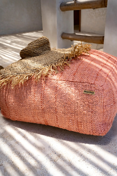 A hand-dyed cotton pouffe graces the sunlit patio, adorned with a straw hat. In the background, wooden steps cast playful shadows on the floor, adding charm to this delightful home accessory setup.