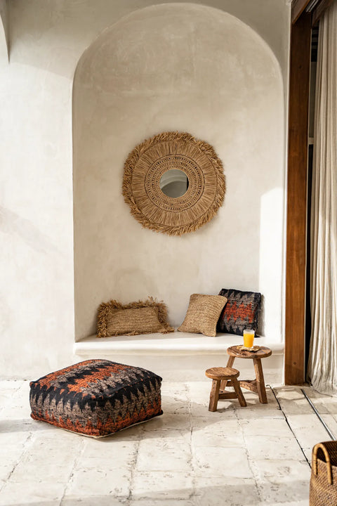 A cozy alcove features a round woven mirror on the wall, patterned cushions on a built-in bench, and an upcycled teak wood stool with a glass of orange juice. A large pouf rests on the stone floor beneath a soft curtain.