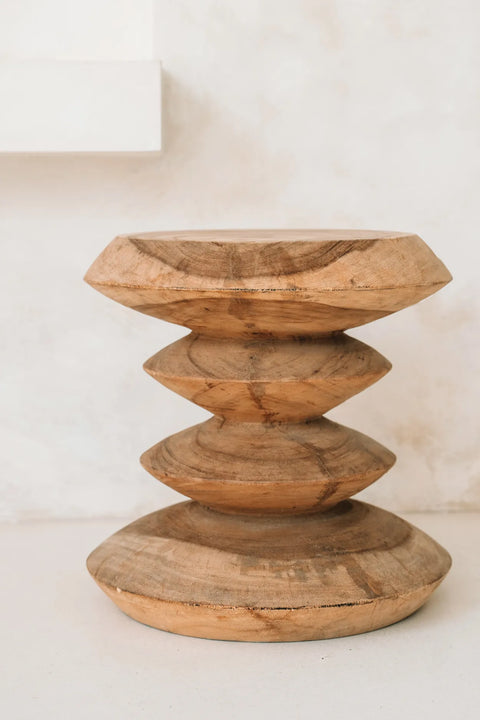 A handmade side table with a unique, sculptural design features three stacked, disk-like shapes with rounded edges. Crafted from teak wood and set against a neutral background, it exudes a natural summer vibe.