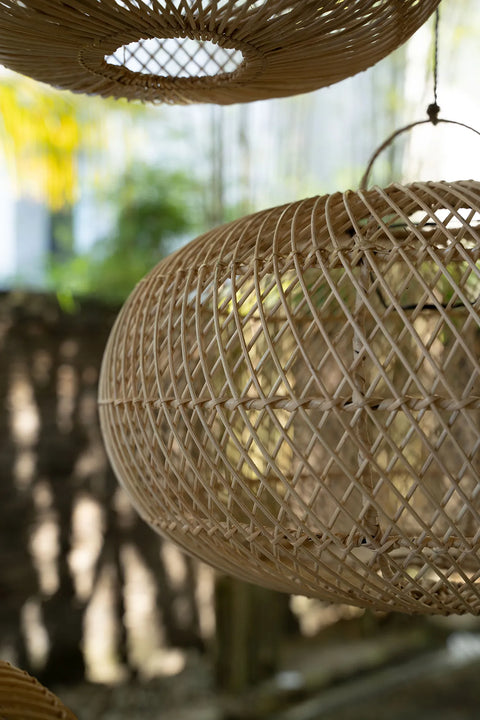 Close-up of a woven rattan pendant light hanging outdoors with sunlight filtering through. Soft-focus foliage and a stone wall are visible in the background, creating a natural and warm ambiance.