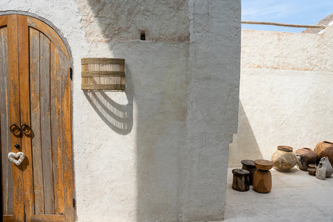 In a rustic outdoor setting, a wooden door with a heart decoration complements a rattan wall applique casting intricate shadows. An array of ceramic pots and wooden stools on the light-colored stone floor enhances the coastal vibe.