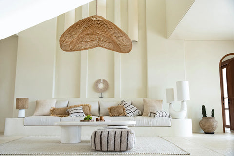 A modern living room featuring a white sofa with assorted textured cushions, a large handcrafted rattan pendant lamp, a minimalist low table, and a striped pouf. The space is adorned with a neutral color palette and boasts a tall, narrow decorative wall feature.