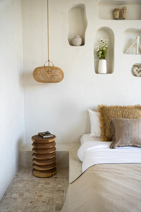 A minimalist bedroom with a neutral color palette. Features include a bed with beige and white linens, a textured pillow, wall shelves with decorative items, a round wooden side table, and a rattan sphere pendant light casting enchanting ceiling shadows.