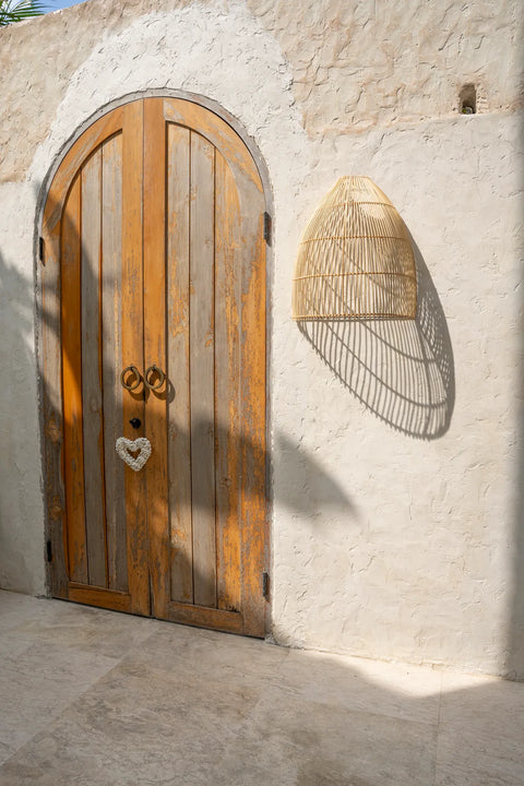 A rustic wooden door with an arched top features a heart-shaped decoration. A rattan light fixture casts a patterned shadow on the white textured wall in bright sunlight, creating an enchanting spectacle.