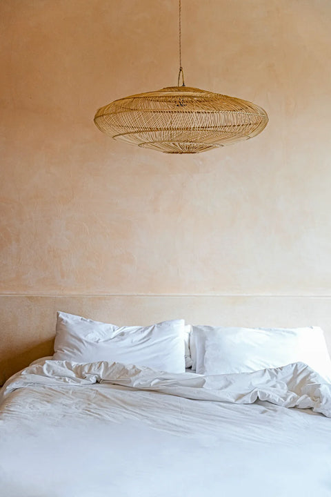 A minimalist bedroom features a large bed with white linens and pillows. A woven rattan pendant lamp hangs above, adding texture to the soft beige walls that create a warm and cozy atmosphere.