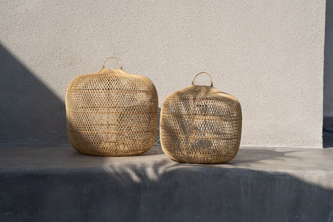 Two woven wicker baskets with handles sit on a concrete ledge, casting intricate shadows on the surface. The background is a plain light-colored wall, evoking the aesthetic of rattan art, while sunlight creates a soft, warm atmosphere.