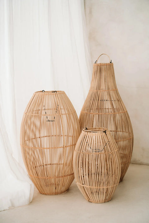 Three tall, wicker baskets of varying sizes stand on a light floor against a backdrop of soft, white fabric. The baskets are cylindrical with a natural, textured appearance that complements the minimalist setting, much like a rattan pendant light casting gentle shadows.