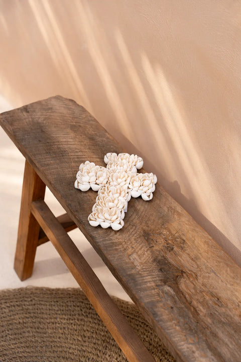 A decorative coastal cross made of white shells rests on a rustic wooden bench under soft lighting. Shadows from the sun create a warm, serene atmosphere, enhancing this coastal decoration. A textured mat is partially visible on the floor.